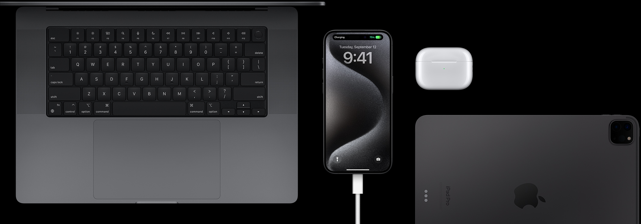 „iPhone 15 Pro“ su į jį įkištu USB-C laidu, apsuptas „MacBook Pro“, „AirPods Pro“ ir „iPad“