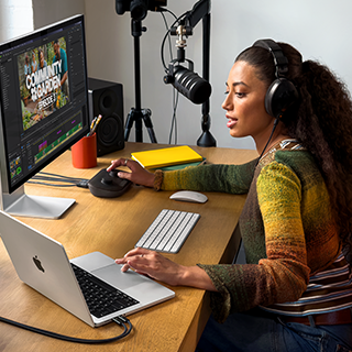 Žena sedí doma u pracovního stolu a používá MacBook Pro připojený k externímu monitoru, má nasazená sluchátka a mluví do mikrofonu