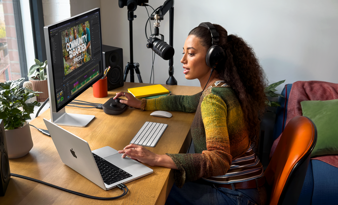 Evindeki çalışma masasında harici bir ekrana bağlı MacBook Pro ile çalışan bir kadın, kulaklık takıyor ve bir mikrofona konuşuyor