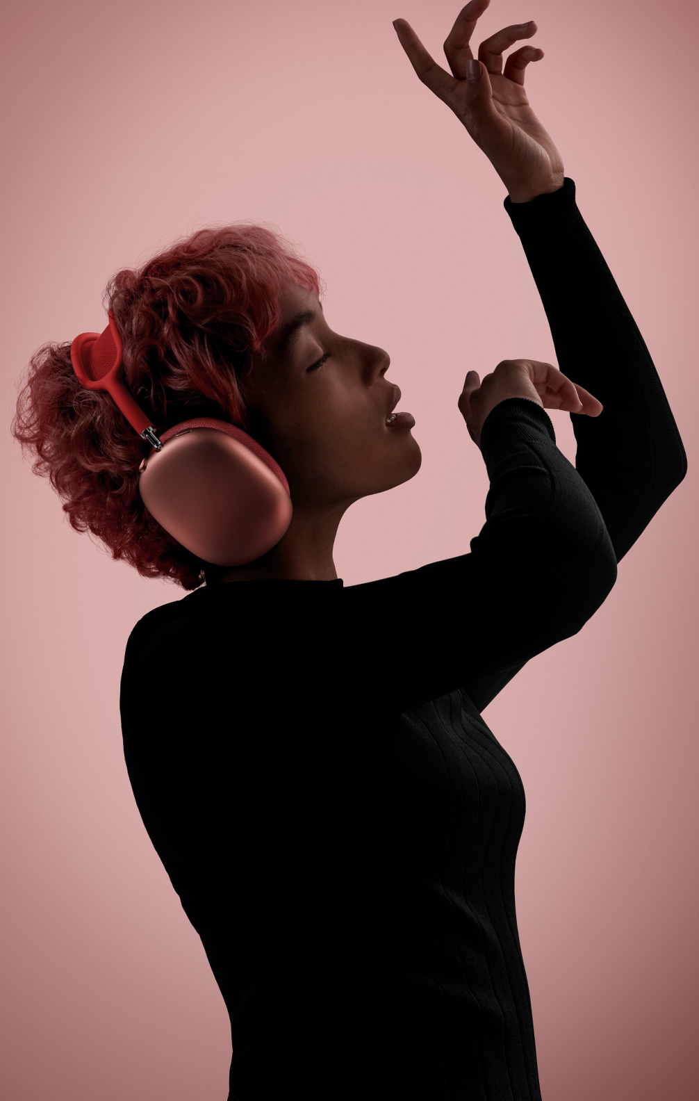Sivuprofiili pinkkejä AirPods Max ‑kuulokkeita käyttävästä henkilöstä, jolla on silmät kiinni ja joka liikehtii kädet ilmaan nostettuina.