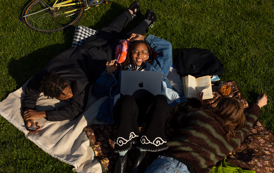 三名大学生躺在公园里的毯子上。一名学生有一部iPhone。一个学生有MacBook Air和苹果耳机。一个学生有一本平装书。