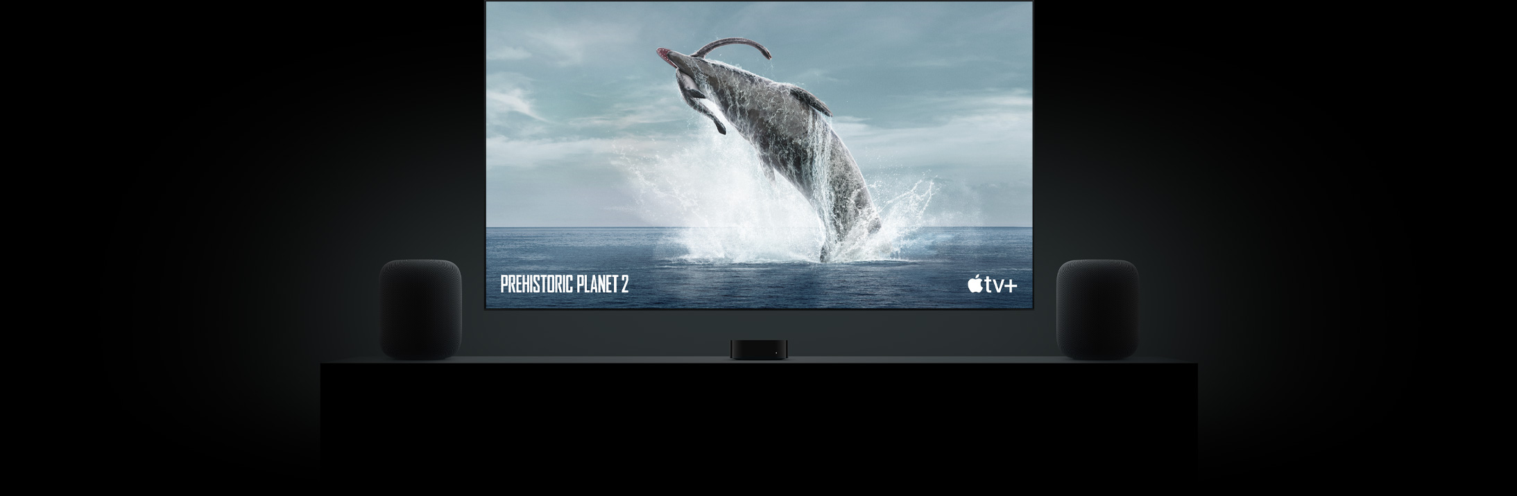 Stor flatskjerm-TV med et naturtro bilde av en dinosaur fra Prehistoric Planet. TV-en henger over en Apple TV og er omgitt av to HomePod-høyttalere på et konsollbord i stuen