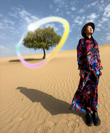 マルチカラーのワンピースを着た女性が、砂漠に立っている写真。背景には1本の木があり、光り輝く虹色のグラデーションの円で囲まれている。背景に写っているものを消すApple Intelligenceの機能を示している