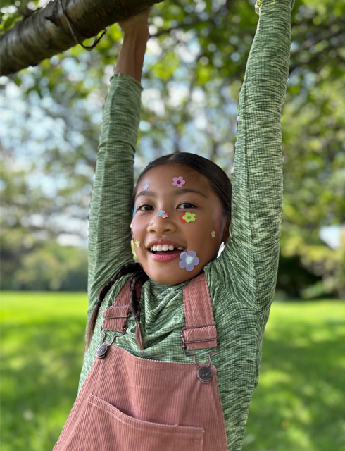 Nærbilde av en jente med blomsterklistremerker på ansiktet, tatt med 2x tele av optisk kvalitet på iPhone 15
