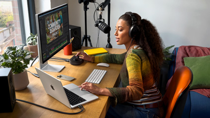 Una donna che lavora alla scrivania di casa con un MacBook Pro collegato a due monitor esterni, indossa un paio di cuffie e parla in un microfono