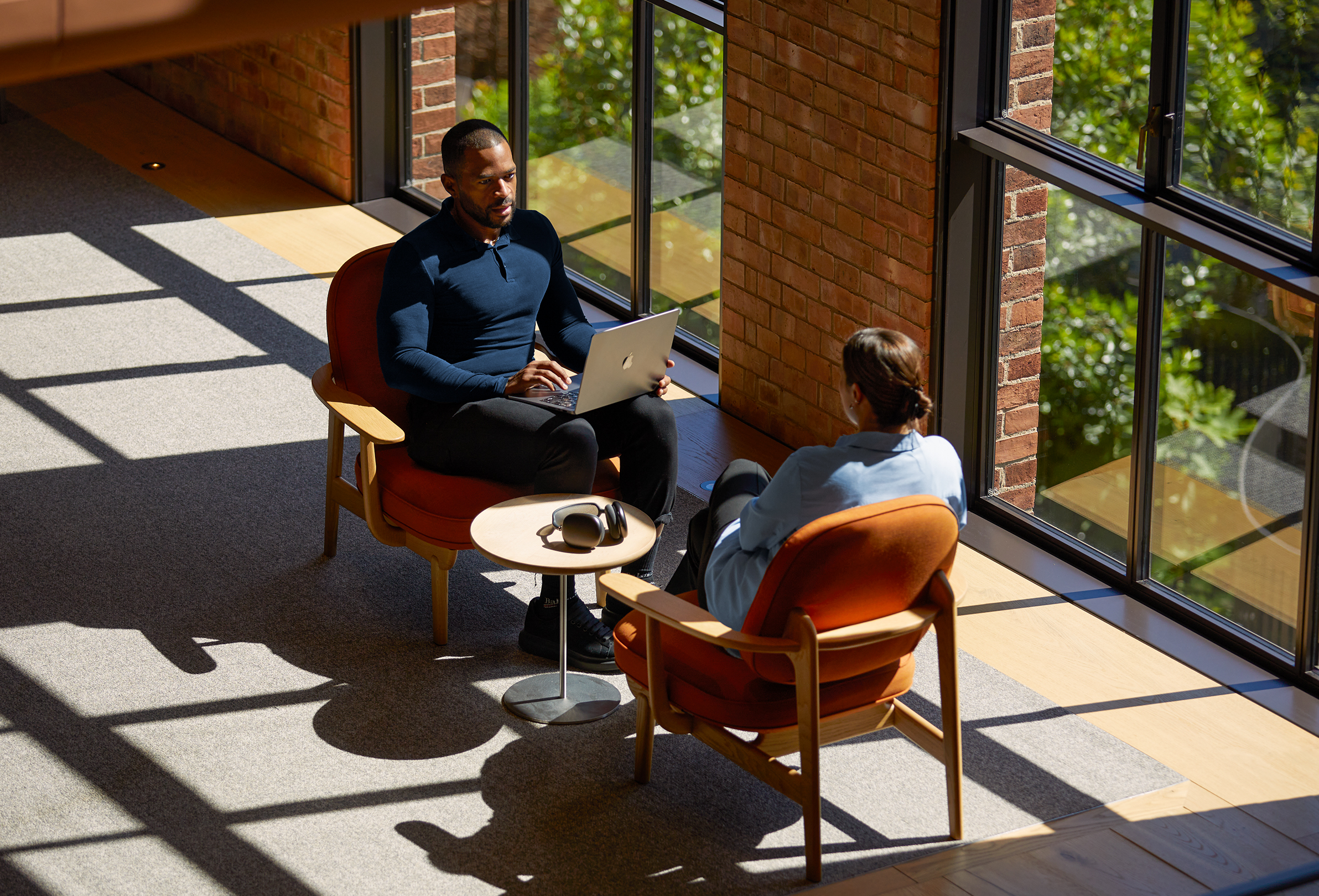 Two Apple colleagues seated in armchairs, talking to each other while one uses a MacBook.