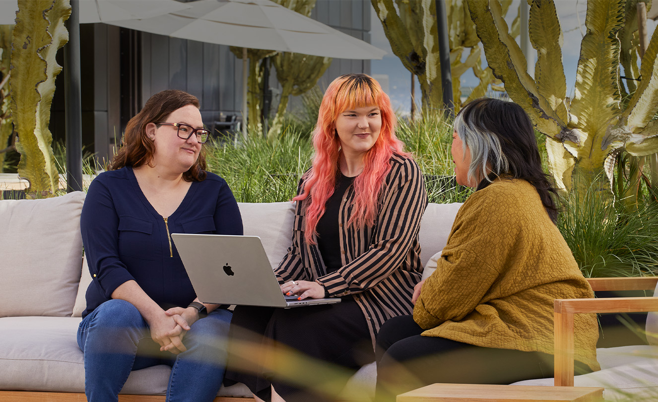 Tři zaměstnankyně společnosti Apple, jedna z nich s MacBookem, sedí společně venku na terase.
