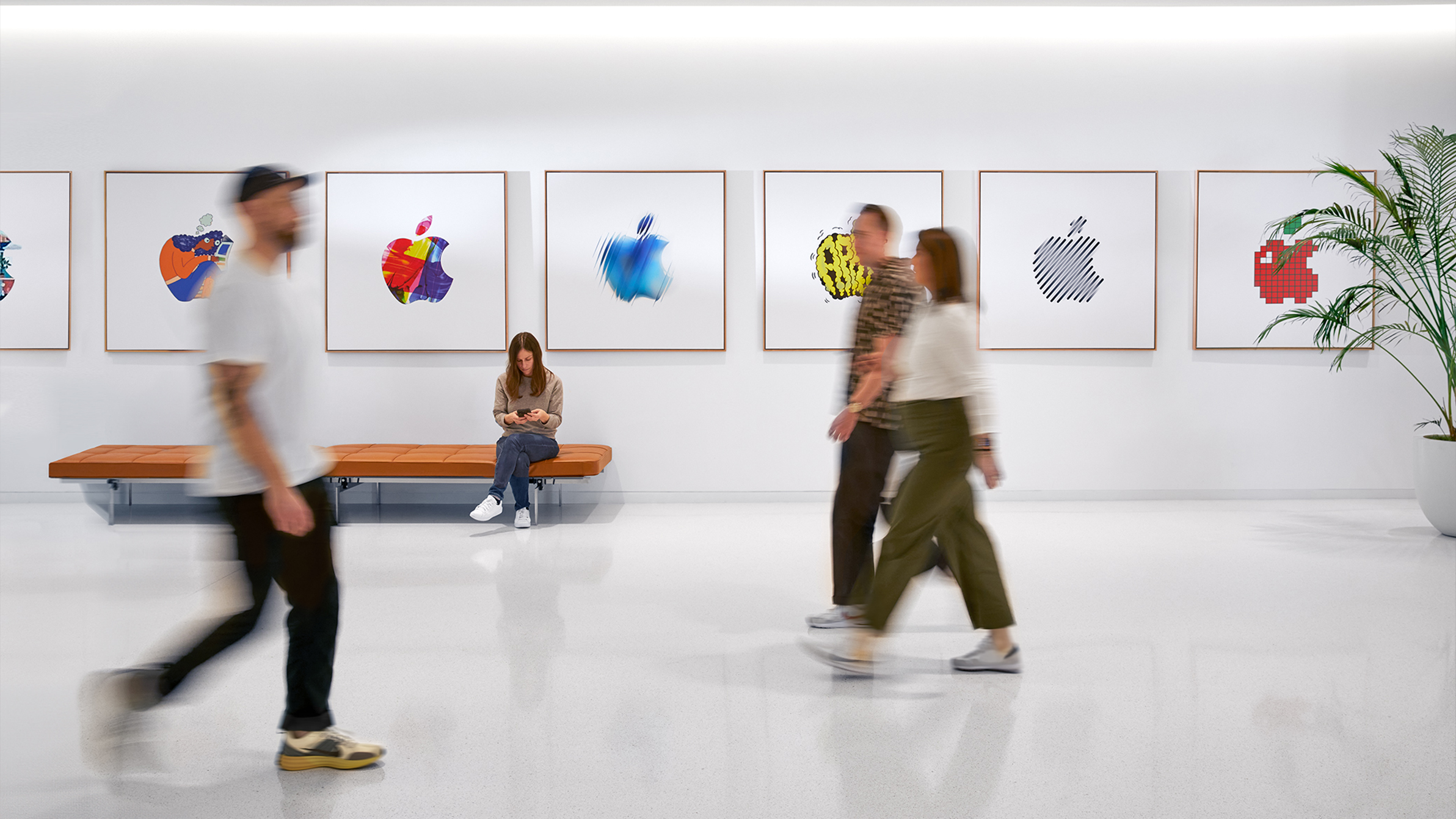 Tři zaměstnanci Apple procházejí v interiéru kolem stěny s barevnými logy Apple a jedna zaměstnankyně sedí na lavičce.