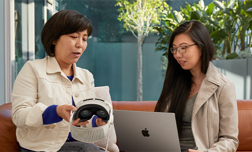 Jessica používá MacBook v místnosti s otevřenými dveřmi na terasu a vedle ní sedí její kolegyně, která jí ukazuje Apple Vision Pro.