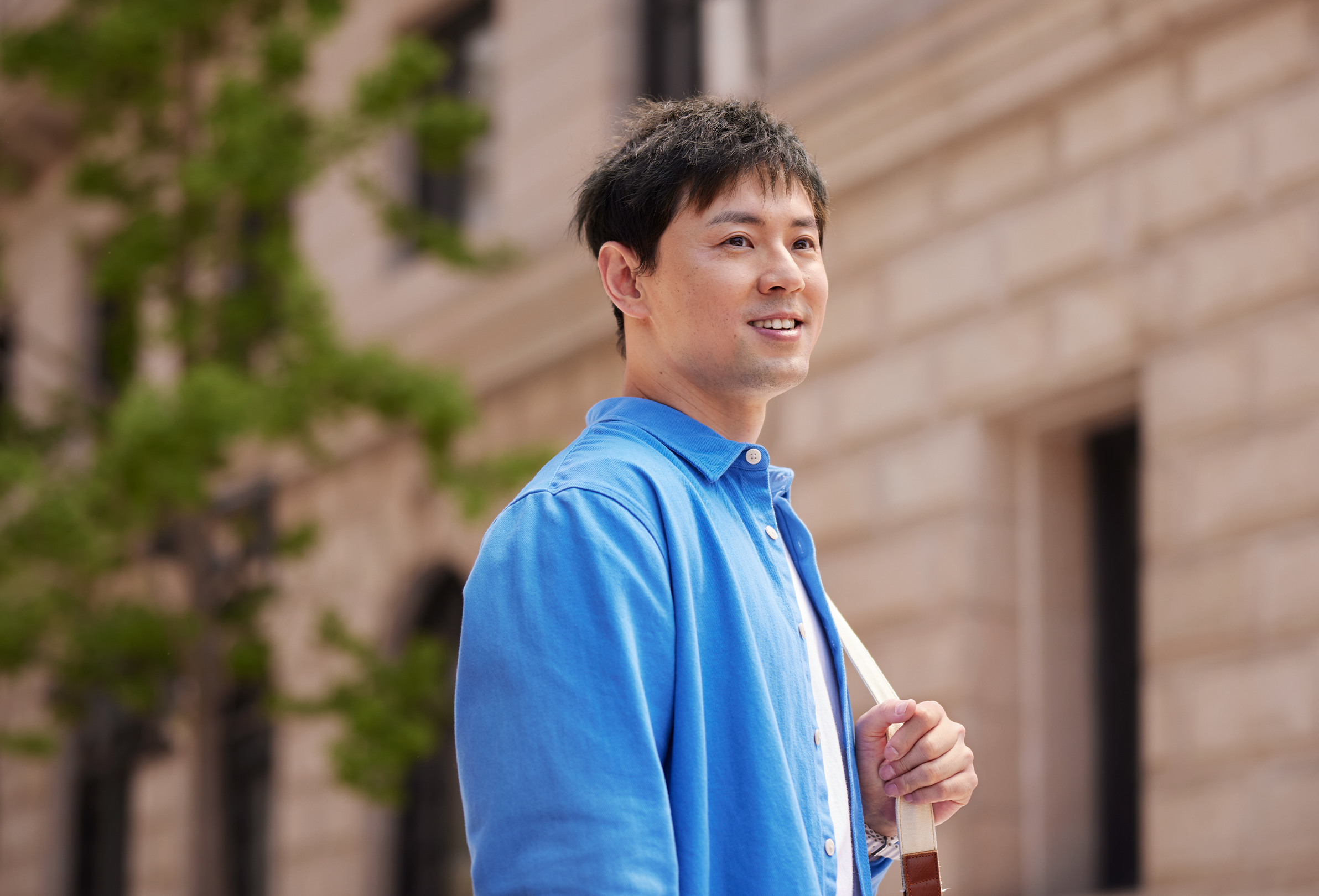 Ein Apple Teammitglied steht lächelnd vor einem Bürogebäude aus Backsteinen. Es trägt eine Schultertasche.