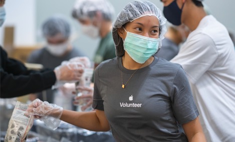 一位穿著 Apple 志工 T 恤的 Apple 實習生在志工活動中包裝物品，微笑著看向旁邊。