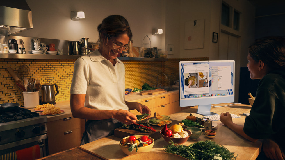 圖片展示一位家長和孩子在廚房裡邊煮飯邊使用搭載 M3 的藍色新款 iMac 以及與之色彩搭配的鍵盤和滑鼠。