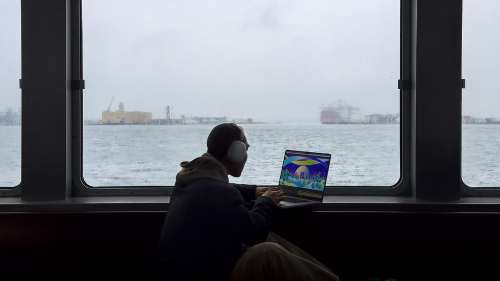 Una persona con audífonos usando la nueva MacBook Pro mientras mira por la ventana hacia un puerto.