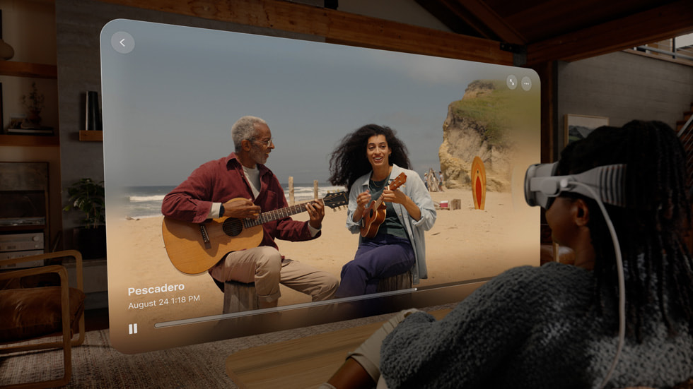 Una persona en su sala con un Apple Vision Pro reproduce un video espacial en una ventana que muestra dos personas que tocan música en una playa.