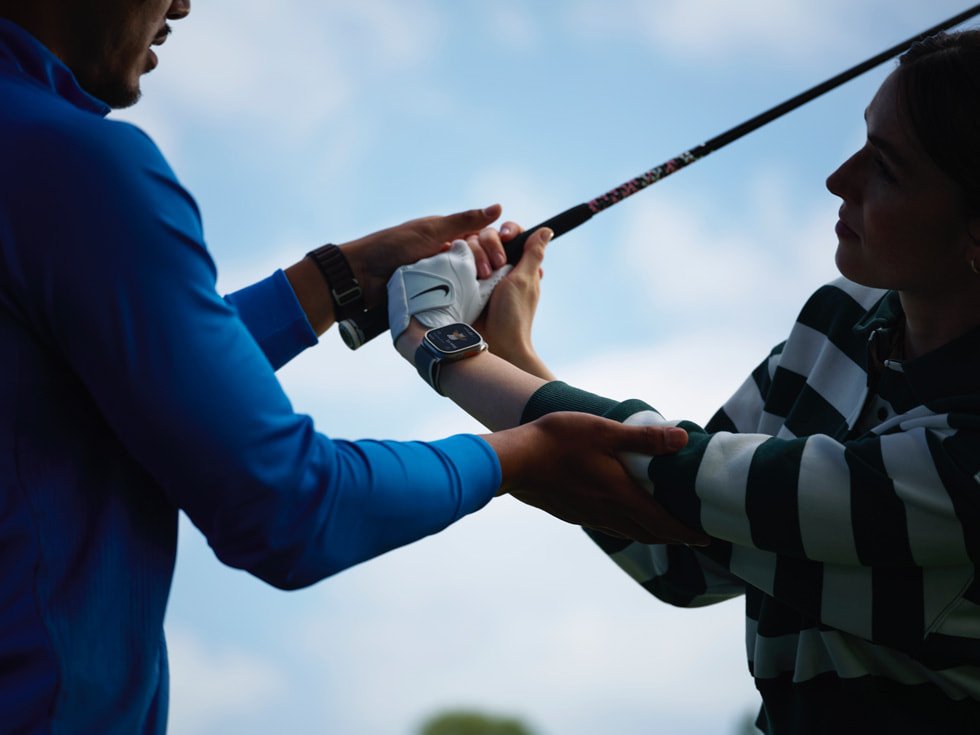 Ilustracja przedstawiająca instruktora gry w golfa, który pomaga uczącej się osobie wykonać zamach kijem golfowym. Osoba ucząca się ma założony Apple Watch.