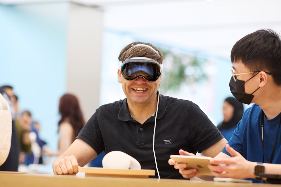 A customer smiles while wearing Apple Vision Pro as a team member looks on.