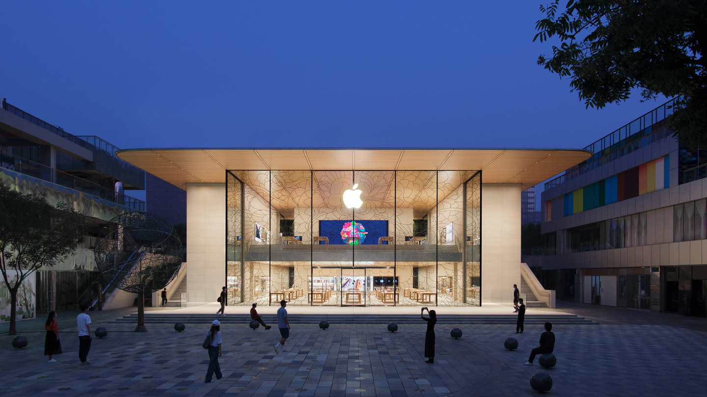 Una vista exterior del frente de la tienda Apple Sanlitun al anochecer muestra el interior iluminado que enfatiza la transparencia y el flujo del diseño del edificio. 