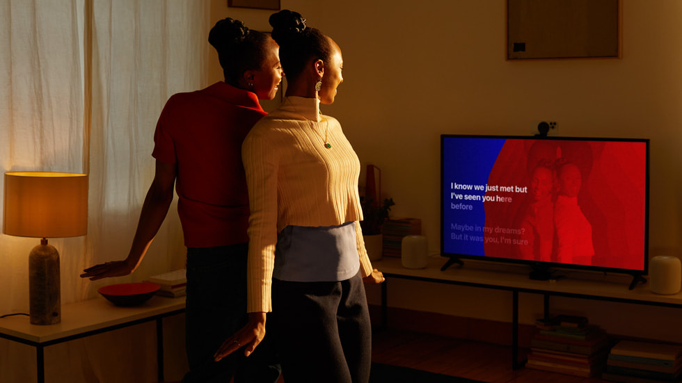 Deux utilisatrices Apple se tiennent dos à dos dans leur salon et apparaissent également sur l’écran de leur télévision.
