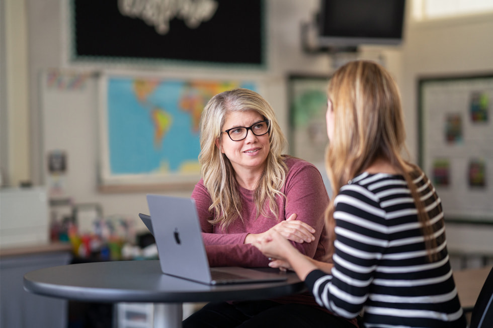 Downey Unified 教學科技教練 Marnie Luevano 在教室裡與一位沒有面對鏡頭的成年人交談，桌上擺著 MacBook Air。