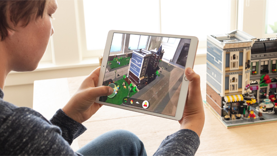 Jongen die in een kamer voor een gebouw van LEGO zit, met in zijn handen een iPad met LEGO AR City op het scherm. 