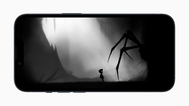 En iPhone 14 med et stillbilde fra Playdead’s LIMBO+ som viser en enorm edderkopp som lurer i skyggene nær et lite barn.
