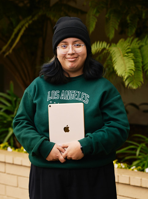 Un portrait d’Angela Ibarra, élève de l’école Exceptional Minds. Angela porte un sweat vert sur lequel est écrit « Los Angeles, California », des lunettes de vue et un bonnet noir.