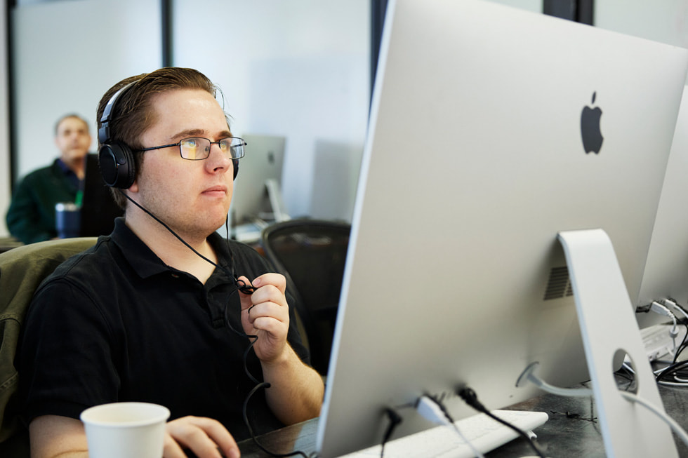 Matthew Rohde, student bij Exceptional Minds, die werkt op een Mac in een lokaal op de campus. Rohde draagt een zwart poloshirt en een koptelefoon.