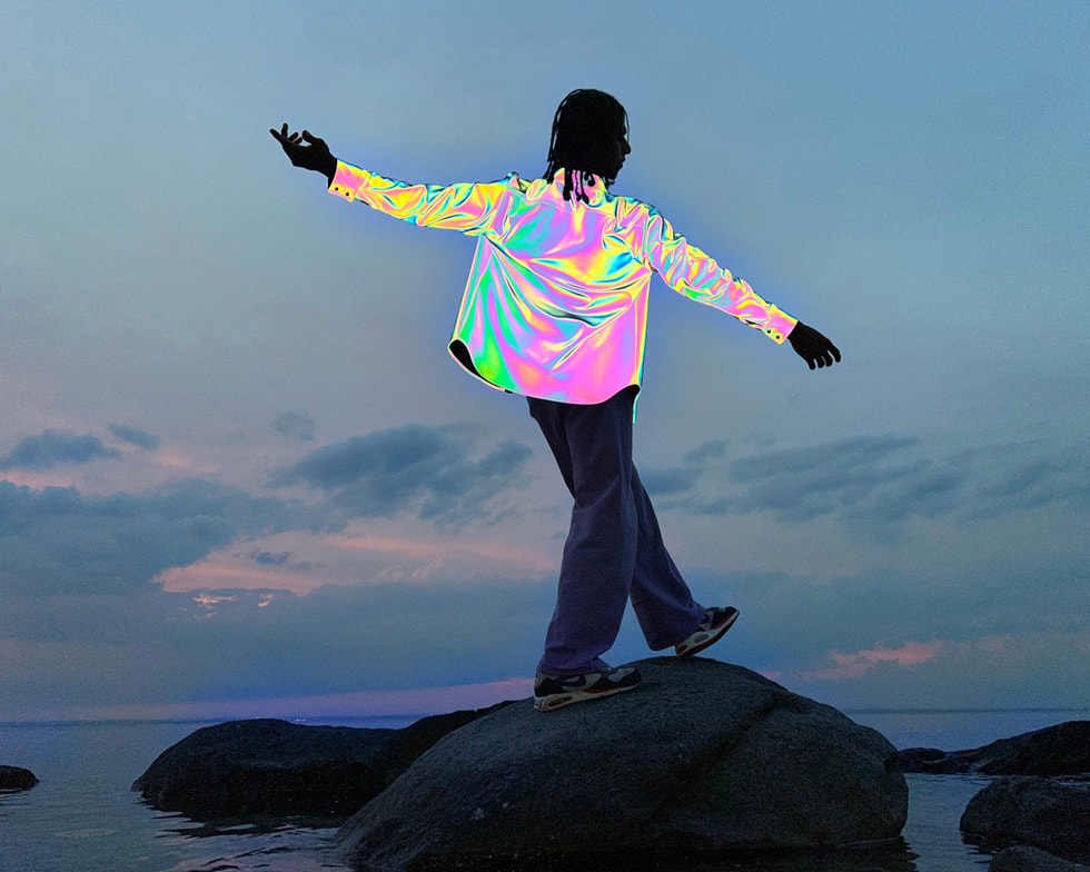 ในภาพนี้ซึ่งถ่ายด้วย iPhone เป็นภาพบุคคลใส่เสื้อเชิ้ตสีเมทัลลิกยืนพิงก้อนหินที่อยู่ในทะเล