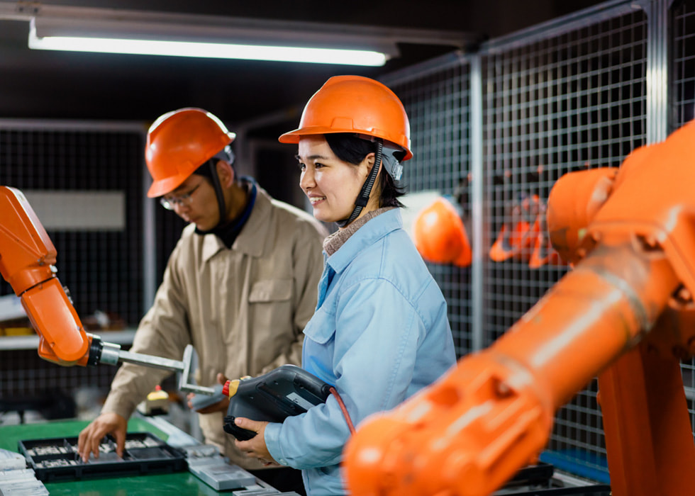 Due persone che lavorano nella filiera e utilizzano un macchinario in una fabbrica indossando caschi protettivi.