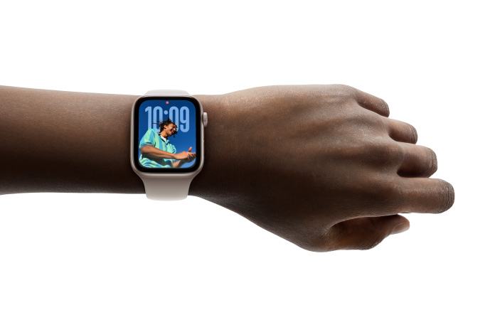 特寫手腕上戴著搭配橡膠錶帶的玫瑰金色 Apple Watch,錶面展示以全新 Liquid Glass 設計呈現的時鐘 app