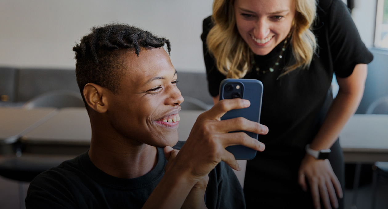 Foto de un joven sonriente con visión reducida que muestra la pantalla de su iPhone por encima de su hombro izquierdo a una mujer que está de pie detrás y también sonríe