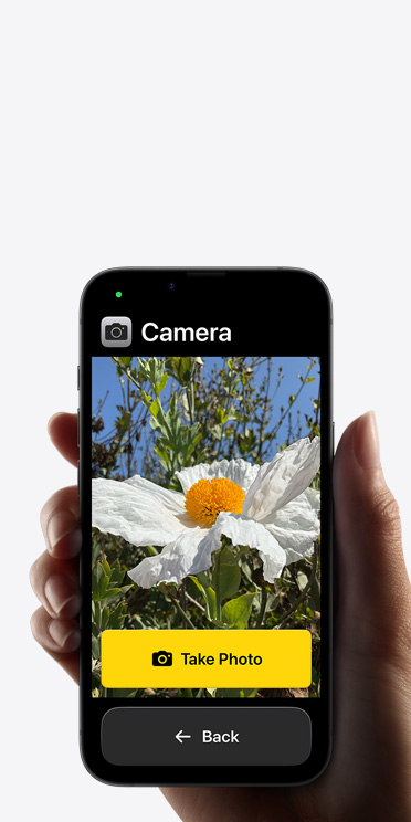 Layar iPhone 16e menampilkan antarmuka aplikasi Kamera yang disederhanakan dengan bunga putih di frame, tombol “Ambil Foto”, dan tombol “ Kembali” di bawah