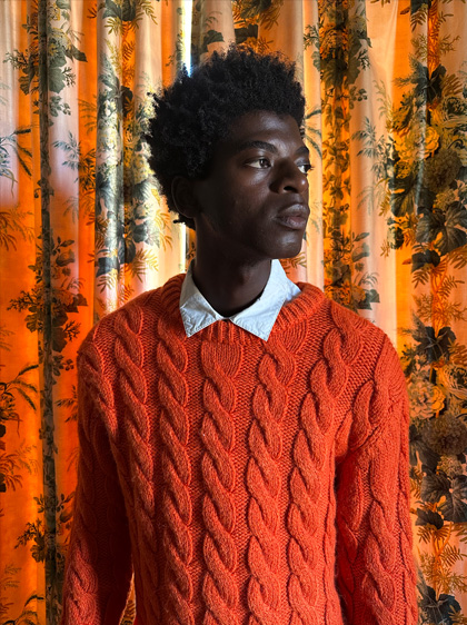 Uma foto de um homem com um suéter vermelho brilhante em frente a cortinas estampadas. A foto foi tirada com pouca luz na câmera principal.