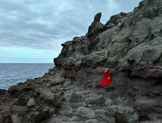 Ein Low-Light-Foto einer Frau in rotem Kleid, die vor einem felsigen Hintergrund posiert, aufgenommen mit der Hauptkamera.